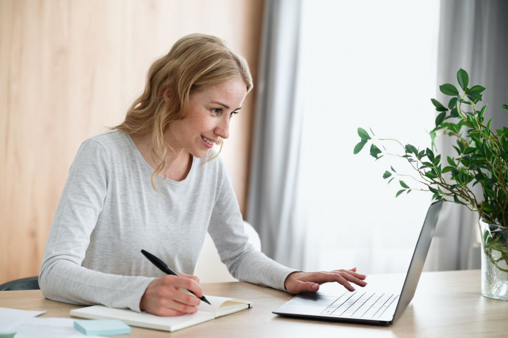 Woman smiling while working on a laptop and taking notes.